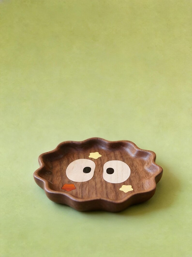 May include: A decorative wooden tray with a wavy edge. The tray is brown with inlaid details forming a cartoon face: large white eyes with black pupils, an orange beak, and yellow star accents. The tray is set against a solid green background.