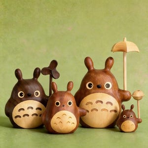 Puede incluir: Colecci&oacute;n de cinco figuras de madera de Totoro de varios tama&ntilde;os. Las figuras son principalmente marrones con detalles de madera m&aacute;s claros en sus vientres. Una sostiene un peque&ntilde;o paraguas y otra una hoja de madera. El fondo es verde liso.