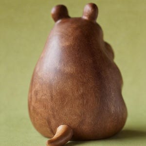 May include: A brown wooden bear figurine, viewed from the back, with two small ears and a curved tail. The bear has a smooth, polished finish and sits on a green surface. The figurine is a decorative item.