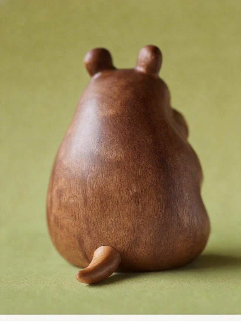 May include: A brown wooden bear figurine, viewed from the back, with two small ears and a curved tail. The bear has a smooth, polished finish and sits on a green surface. The figurine is a decorative item.