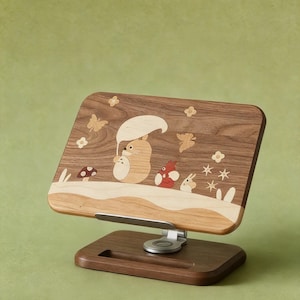 May include: A wooden phone or tablet stand featuring a rectangular back panel with inlaid wood designs of a cartoon creature holding an umbrella, small animals, mushrooms, butterflies, and flowers. The stand has a metal hinge and a dark wood base.