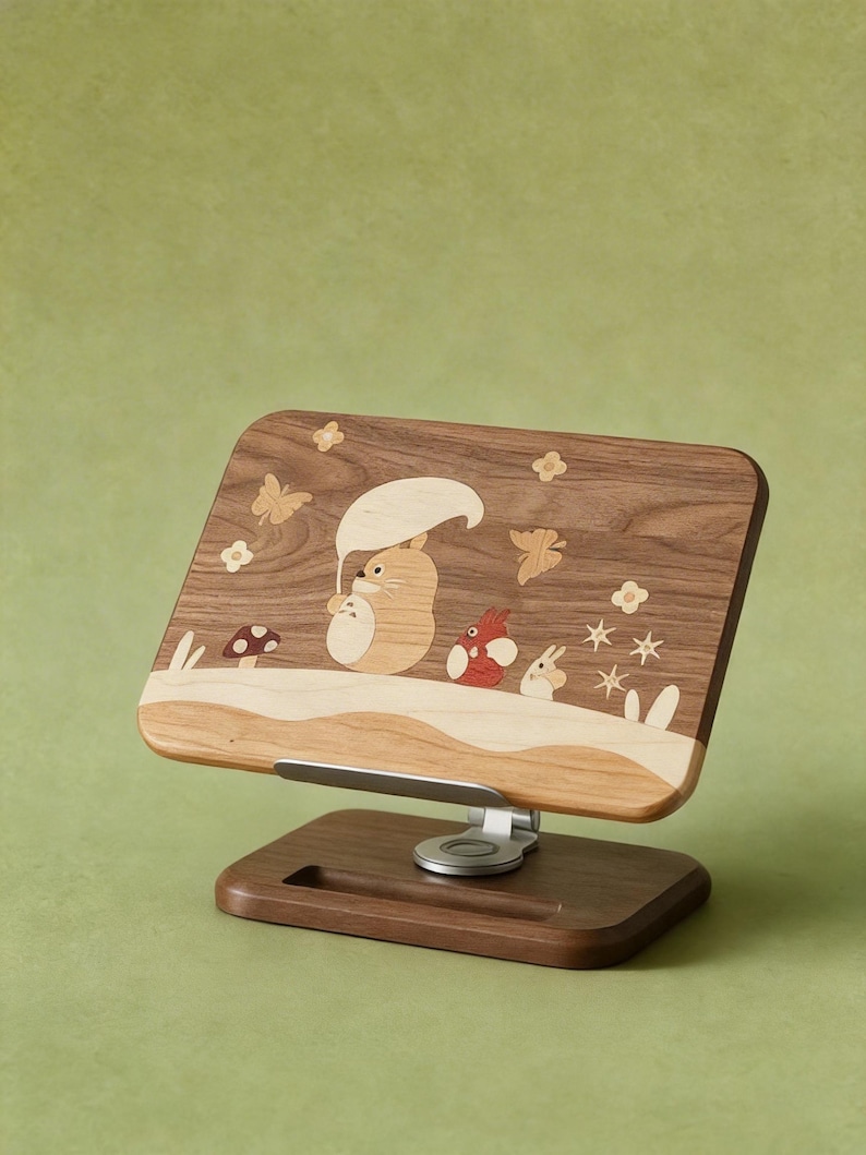 May include: A wooden phone or tablet stand featuring a rectangular back panel with inlaid wood designs of a cartoon creature holding an umbrella, small animals, mushrooms, butterflies, and flowers. The stand has a metal hinge and a dark wood base.