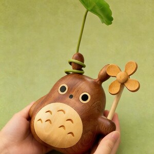 Puede incluir: Figura de madera de un personaje de dibujos animados con vientre marr&oacute;n claro, cuerpo marr&oacute;n oscuro y ojos negros grandes. Una hoja y un tallo verdes brotan de la parte superior. Una flor de madera se sostiene en la mano del personaje.