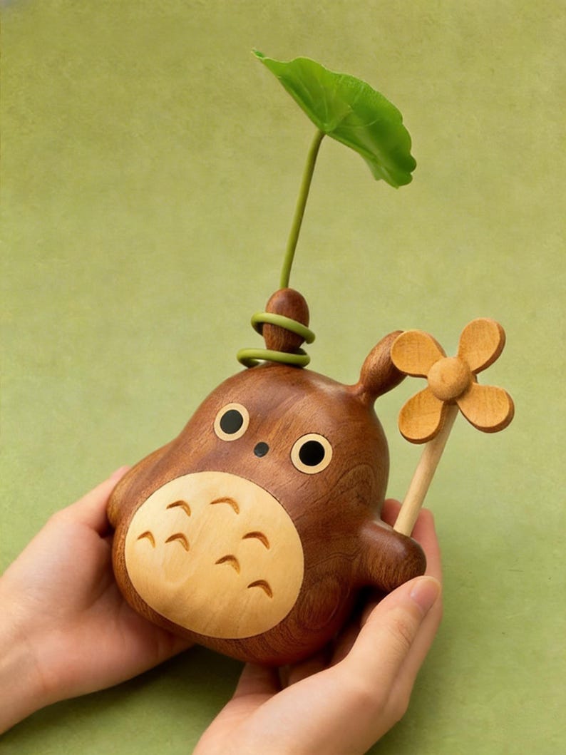 Puede incluir: Figura de madera de un personaje de dibujos animados con vientre marr&oacute;n claro, cuerpo marr&oacute;n oscuro y ojos negros grandes. Una hoja y un tallo verdes brotan de la parte superior. Una flor de madera se sostiene en la mano del personaje.