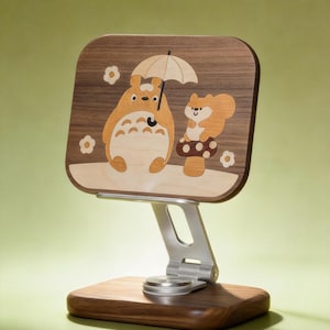 May include: A wooden phone stand featuring a rectangular panel with an inlaid design of a cartoon creature holding an umbrella next to a squirrel on a mushroom. The stand has a silver metal adjustable arm and a solid dark wood rectangular base.