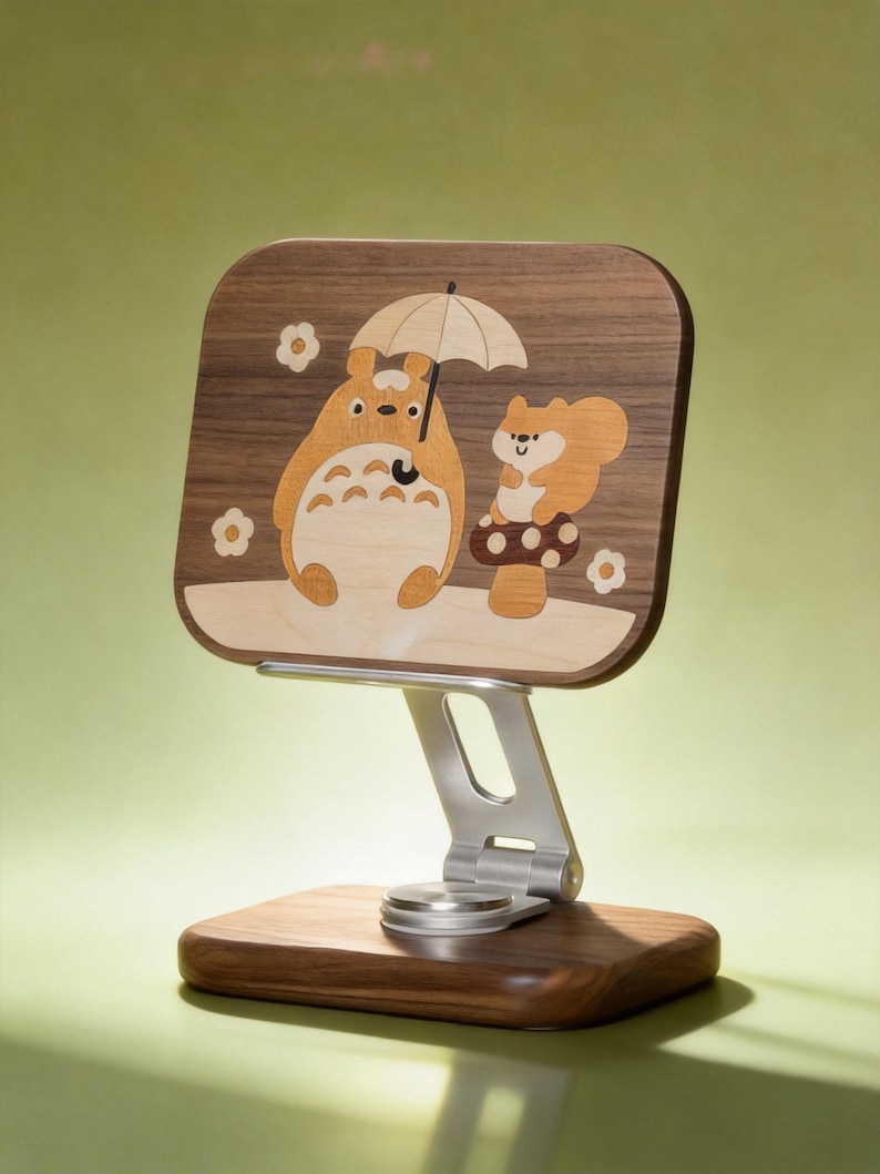 May include: A wooden phone stand featuring a rectangular panel with an inlaid design of a cartoon creature holding an umbrella next to a squirrel on a mushroom. The stand has a silver metal adjustable arm and a solid dark wood rectangular base.
