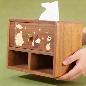 K&ouml;nnte beinhalten: Holz-Taschentuchbox mit einer Schublade und zwei unteren F&auml;chern. Die Box ist mit Cartoon-Hasen- und Sterndesigns verziert. Ein goldfarbener Knopf befindet sich an der Schublade. Wei&szlig;e Taschent&uuml;cher sind oben sichtbar.