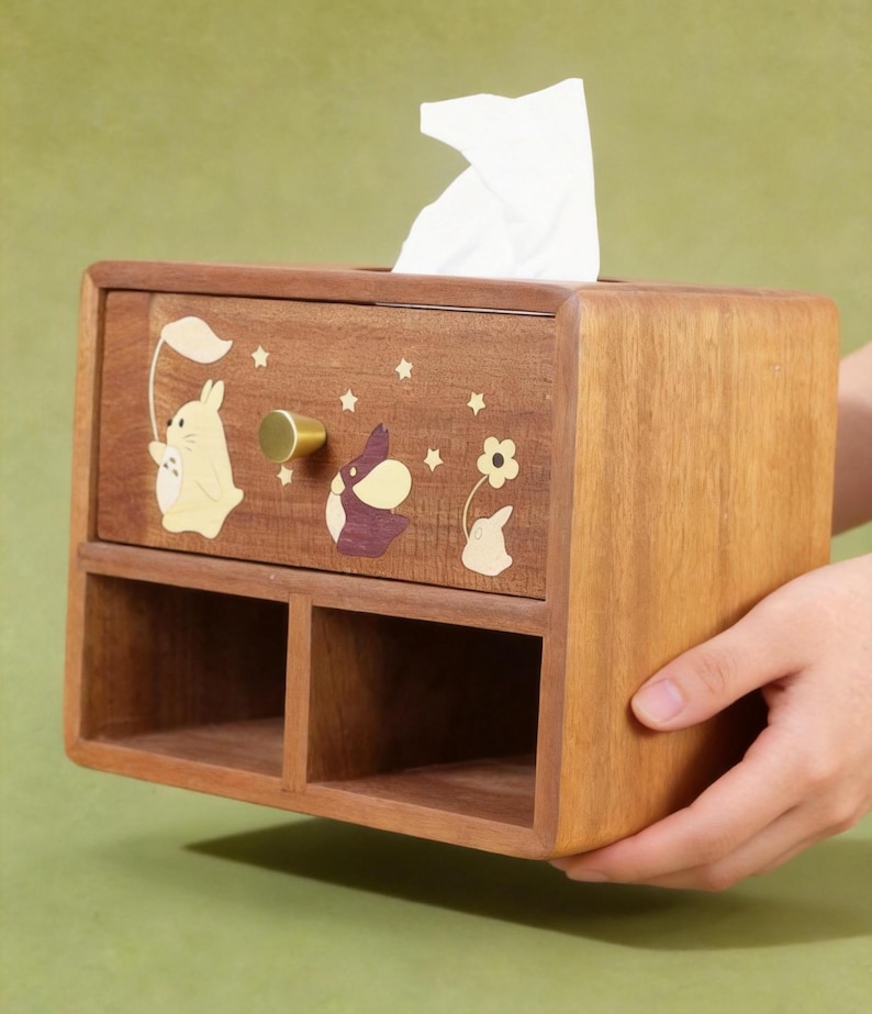 K&ouml;nnte beinhalten: Holz-Taschentuchbox mit einer Schublade und zwei unteren F&auml;chern. Die Box ist mit Cartoon-Hasen- und Sterndesigns verziert. Ein goldfarbener Knopf befindet sich an der Schublade. Wei&szlig;e Taschent&uuml;cher sind oben sichtbar.