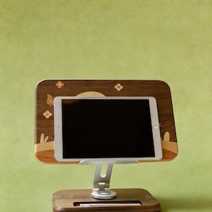 May include: A tablet mounted on an adjustable silver metal stand with a dark wood back panel featuring light wood inlays of flowers and a butterfly. The base is made of dark wood with a horizontal slot containing a white stylus pen.