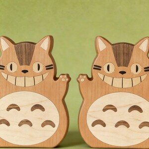 May include: Two wooden cat-shaped figures with smiling faces and outstretched paws. The figures are made of light and dark brown wood, with a light-colored belly and a dark brown stripe on the head. The background is a solid green color.