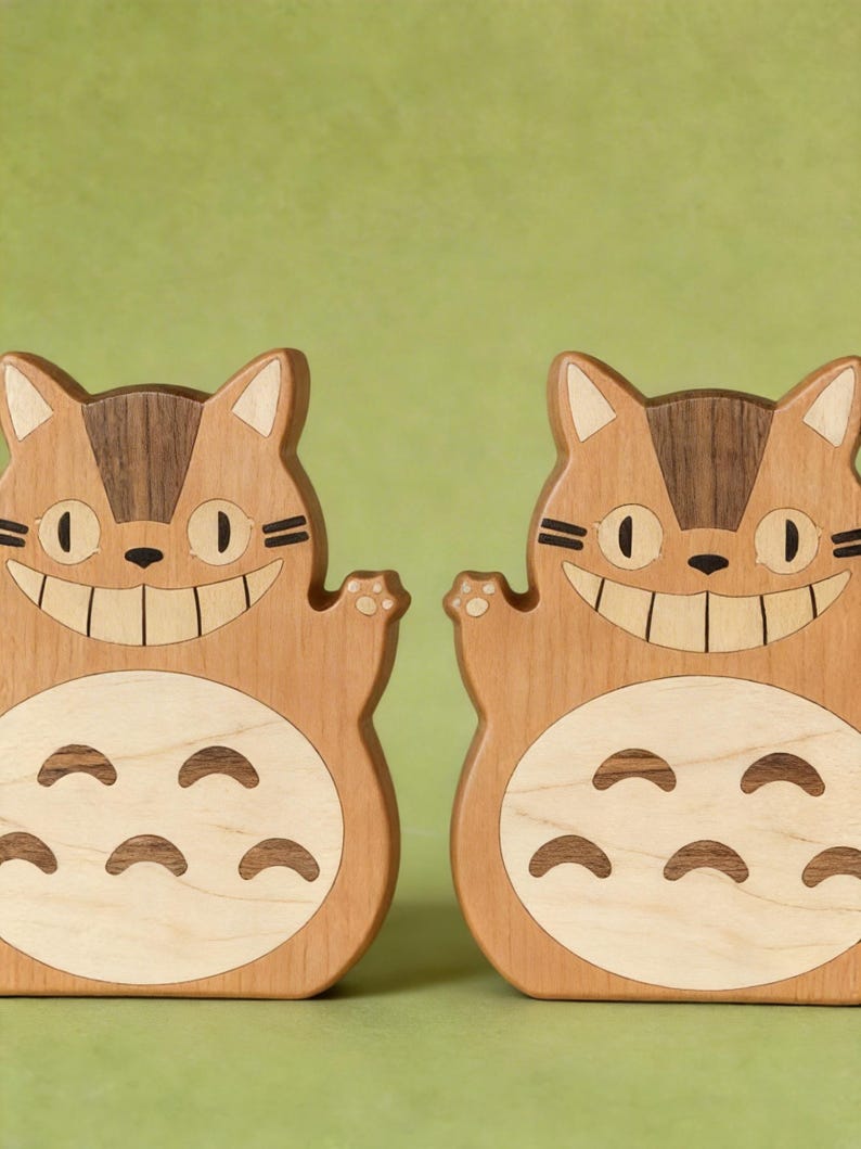 May include: Two wooden cat-shaped figures with smiling faces and outstretched paws. The figures are made of light and dark brown wood, with a light-colored belly and a dark brown stripe on the head. The background is a solid green color.