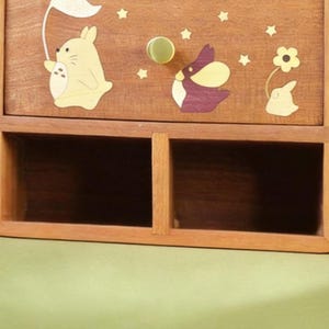 K&ouml;nnte beinhalten: Holz-Aufbewahrungsbox mit zwei offenen F&auml;chern und einer Schublade. Die Schubladenfront ist mit Cartoon-Hasen- und Sterndesigns verziert. Der Schubladengriff ist ein runder, goldfarbener Knopf. Die Box ist aus hellbraunem Holz.