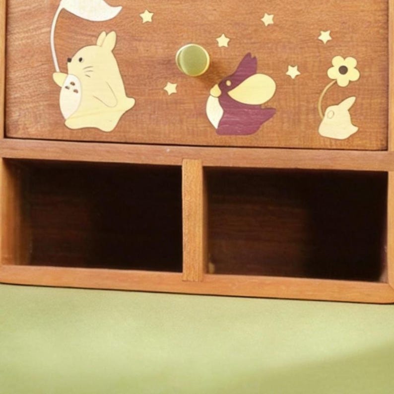 K&ouml;nnte beinhalten: Holz-Aufbewahrungsbox mit zwei offenen F&auml;chern und einer Schublade. Die Schubladenfront ist mit Cartoon-Hasen- und Sterndesigns verziert. Der Schubladengriff ist ein runder, goldfarbener Knopf. Die Box ist aus hellbraunem Holz.