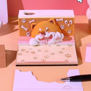 May include: A layered paper notepad featuring a 3D orange cat design. The notepad has a light pink and peach color scheme, with paw print details. A pen rests on a separate sheet of paper.