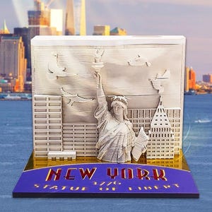 May include: A layered sculpture depicting the Statue of Liberty and New York City buildings. The base features the text "NEW YORK 1776 STATUE OF LIBERTY" in gold on a blue background. A cityscape and water form the backdrop.