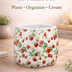 May include: A square, white ceramic 3D puzzle pot with a strawberry design. The pot has a white interior and is decorated with red strawberries, green leaves, and white flowers. Text on the pot reads "3D Puzzle Pot" and "Sweet Berry Garden."
