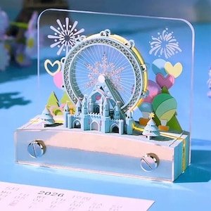 May include: A decorative acrylic display featuring a Ferris wheel, castle, and hot air balloons. The Ferris wheel has a gold rim and a snowflake design in the center. The castle is light blue, and the display includes fireworks and heart-shaped balloons.