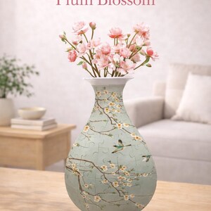 Könnte beinhalten: Eine dekorative Vase mit Blumen- und Vogeldesign, die rosa Blumen enthält. Die Vase hat einen hellblauen Hintergrund mit einem Pflaumenblüten- und Vogelmuster. Der Text "Birds in Plum Blossom" wird oben angezeigt.
