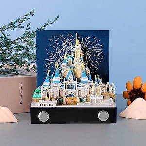 Puede incluir: Modelo 3D detallado de un castillo con detalles azules y dorados, sobre un fondo azul oscuro con fuegos artificiales. El modelo se asienta sobre una base negra con dos botones plateados. Elementos decorativos: una flor y objetos con forma de montaña.