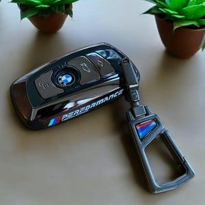 Peut inclure: Une clé de voiture noire avec un logo BMW argenté et les mots "Performance Limited Edition" sur le côté. La clé est attachée à un porte-clés en métal noir avec un logo BMW bleu et blanc.