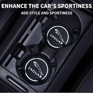 Puede incluir: Posavasos negros para coche con el logotipo blanco de Jaguar. Los posavasos son circulares y encajan en los portavasos del coche. La imagen también incluye el texto "ENHANCE THE CAR'S SPORTINESS" y "ADD STYLE AND SPORTINESS".