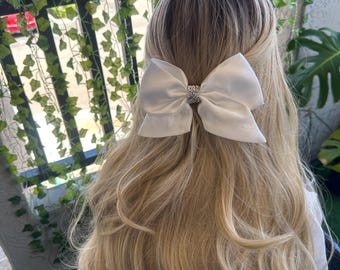 May include: A white satin bow with a rhinestone center, worn in blonde hair.