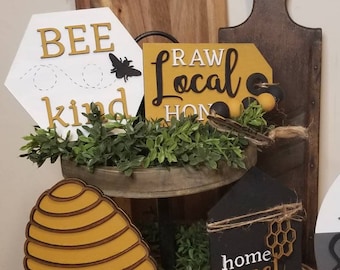 May include: A tiered tray decorated with a beehive, bees, and wooden signs that read "Bee Kind", "Raw Local Honey", and "Home Sweet Hive".