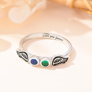 Op de afbeelding: Zilveren ring met twee engelenvleugels aan weerszijden van drie edelstenen. De edelstenen zijn blauw, groen en groen. De ring is gegraveerd met de woorden "Love you forever".