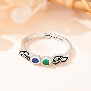 Op de afbeelding: Een zilveren ring met twee engelenvleugels aan weerszijden van drie edelstenen. De edelstenen zijn blauw, groen en groen. De ring is gegraveerd met de woorden "Love you forever".