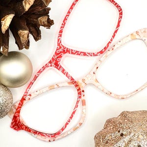May include: Red and white patterned eyeglasses with a double frame design. The glasses have a square shape and are made of plastic.