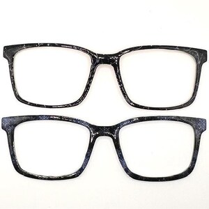 May include: Two pairs of eyeglass frames with a black and white marbled pattern. The frames have a rectangular shape with rounded edges. The lenses are clear, and the frames are set against a white background.