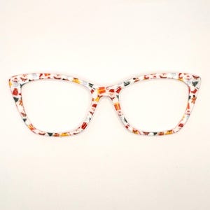 May include: White cat-eye glasses with a colorful pattern of red, yellow, and green. The pattern features small, repeated images of Santa Claus, reindeer, and other Christmas-themed icons.