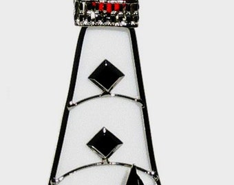 May include: A decorative lighthouse ornament with a red top, white body, and blue base. The lighthouse features black diamond accents and a small black sailboat on the blue water. The ornament is trimmed in silver and has a silver loop for hanging.