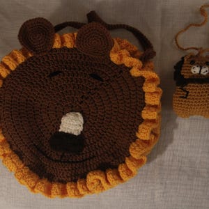 May include: A handmade crochet lion-shaped bag and a matching lion-shaped keychain. The bag is brown with a yellow mane and a smiling face. The keychain is a smaller version of the lion, also in brown and yellow.