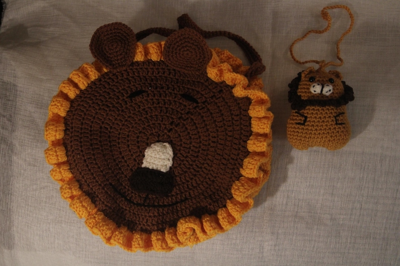 May include: A handmade crochet lion-shaped bag and a matching lion-shaped keychain. The bag is brown with a yellow mane and a smiling face. The keychain is a smaller version of the lion, also in brown and yellow.