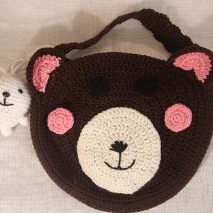 May include: A handmade brown crochet bear-shaped bag with pink accents and a white face. A small white crochet bear charm is attached. The bag has a handle and is likely for children.