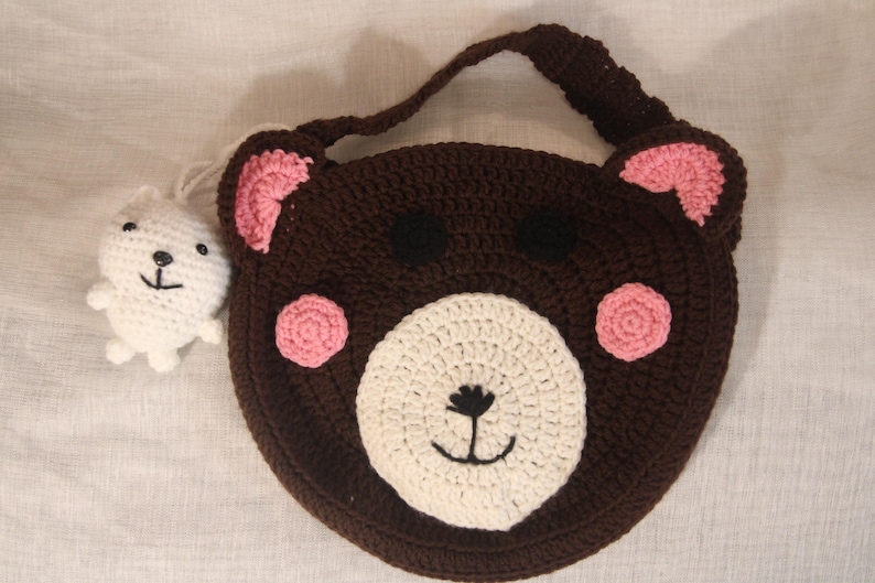 May include: A handmade brown crochet bear-shaped bag with pink accents and a white face. A small white crochet bear charm is attached. The bag has a handle and is likely for children.