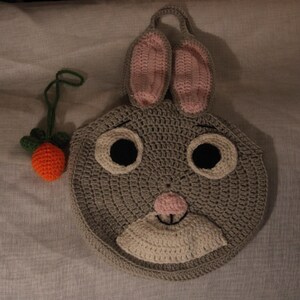 May include: A handmade crochet bunny face in gray, with pink inner ears, black eyes, and a white muzzle. A small orange carrot with green leaves is next to the bunny. The bunny has a loop for hanging.