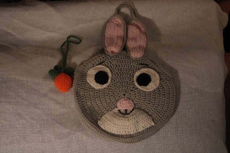 May include: A handmade crochet bunny face in gray, with pink inner ears, black eyes, and a white muzzle. A small orange carrot with green leaves is next to the bunny. The bunny has a loop for hanging.