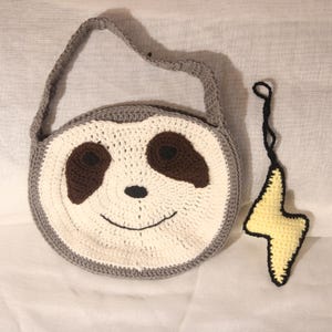 May include: A handmade crochet bag shaped like a sloth's face, featuring a gray border, white and brown details, and a smiling mouth. A yellow lightning bolt charm with a black outline hangs nearby.
