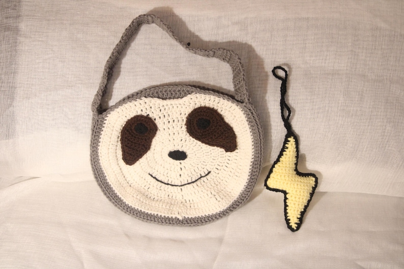 May include: A handmade crochet bag shaped like a sloth's face, featuring a gray border, white and brown details, and a smiling mouth. A yellow lightning bolt charm with a black outline hangs nearby.