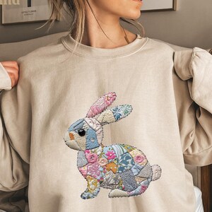 May include: A sand-colored sweatshirt featuring a colorful patchwork bunny design. The bunny is made of various fabric patterns, including floral and geometric prints. The sweatshirt has long sleeves and a crew neck.