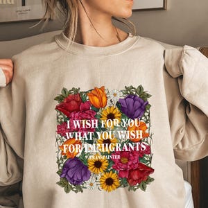 May include: A sand-colored sweatshirt featuring a floral design with the text "I wish for you what you wish for immigrants." The design includes red, orange, purple, and yellow flowers. The sweatshirt is a casual style.
