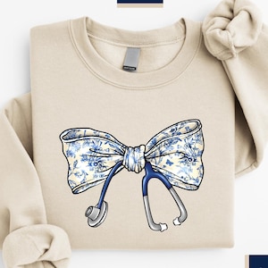 May include: A beige sweatshirt with a blue and white floral bow and a stethoscope. The text 'KS khanshirtus' is in the top left corner of the image. The text 'SAND' is in the bottom right corner of the image.