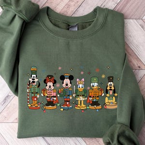 May include: Military green sweatshirt featuring a colorful graphic of Disney characters dressed as nutcrackers. The design includes Mickey Mouse, Minnie Mouse, Donald Duck, Goofy, and others. The sweatshirt has long sleeves and a crew neck.