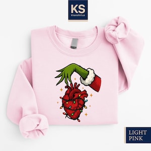 A pink sweatshirt with a graphic design depicting a hand holding a lit heart, surrounded by Christmas lights.