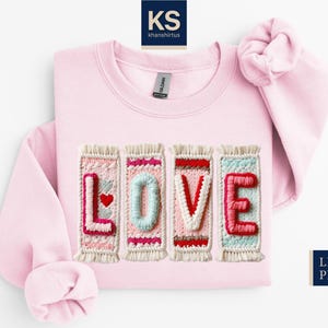 May include: Light pink sweatshirt with the word "LOVE" embroidered across the front. The letters are in a colorful, textured design with a heart accent. The sweatshirt has a crew neck and long sleeves.