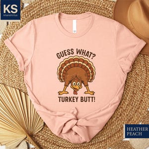 A pink t-shirt with a humorous text and graphic of a turkey, along with a straw hat and a fan, all arranged on a woven mat.
