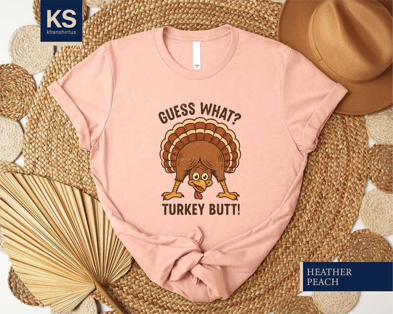 A pink t-shirt with a humorous text and graphic of a turkey, along with a straw hat and a fan, all arranged on a woven mat.