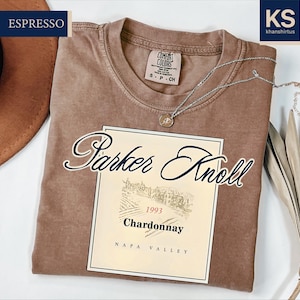 a brown t-shirt with a label that reads "Parker Knoll 1994 Chardonnay Napa Valley" and a small tag that says "Espresso". The t-shirt is laid out on a white surface, with a brown hat and some leaves visible in the background.